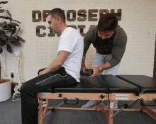 two men are working on a man 's back on a table in front of a brick wall with the word osteopath on it