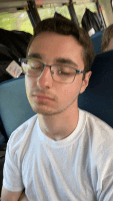 a young man wearing glasses and a white shirt is sitting on a bus with his eyes closed