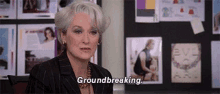 a woman with gray hair is sitting at a desk in front of a bulletin board and says `` groundbreaking '' .