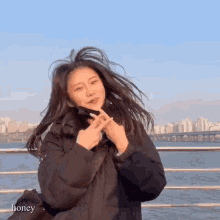 a woman in a black jacket is making a heart shape with her hands while standing in front of a body of water .