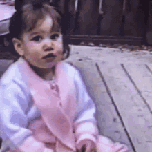 a little girl in a pink and white dress is sitting on a wooden deck making a funny face .