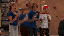 a group of children are standing in front of a climbing wall . one of the children is wearing a shirt that says get sweaty