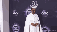 a man wearing a white cape and a polka dot hat stands on a purple carpet at the american music awards