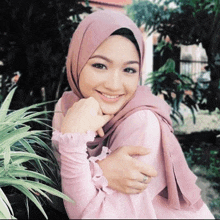 a woman wearing a pink hijab and a pink shirt is smiling and hugging a plant .