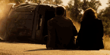 a man and a woman are sitting on the side of a road looking at a car that has turned over