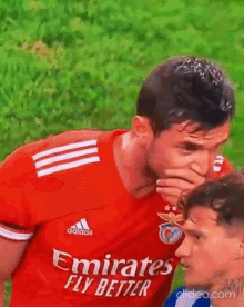 a soccer player wearing a red emirates fly better jersey is covering his mouth while another player looks on .