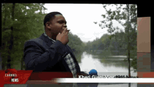 a man in a suit smoking a cigarette in front of a river with channel news written on the bottom of the screen