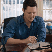 a man in a blue shirt is sitting at a desk writing on a piece of paper with schitts creek written on the bottom