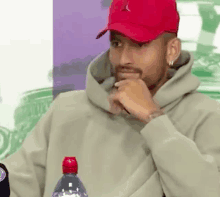 a man wearing a red hat and a hoodie is sitting at a table with his hand on his chin .