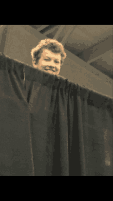 a young man stands behind a black curtain and smiles at the camera