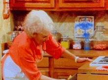 a woman in a red shirt and white apron is standing in a kitchen with a clock on the wall that says trendiest