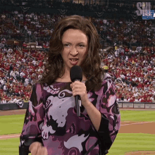 a woman in a purple shirt is holding a microphone in front of a crowd that is watching a baseball game