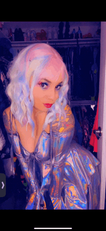 a woman with pink and blue hair is wearing a holographic dress and standing in a closet .