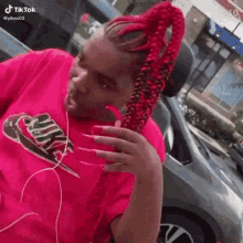 a woman with red braids and long nails wearing a pink nike shirt is standing in front of a car .