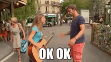 a woman holding a guitar talks to a man on a street with the words ok ok on the bottom