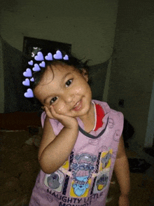 a little girl wearing a purple shirt is smiling with her hand on her face .