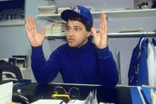 a man in a blue hat and sweater is sitting at a desk with his hands in the air .