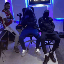 three men are sitting in chairs in a dark room . one of the men is wearing a red and white adidas shirt .