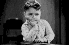 a black and white photo of a young boy sitting at a table with his hand on his chin .