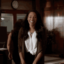 a woman in a white shirt and brown cardigan smiles in front of a clock on the wall