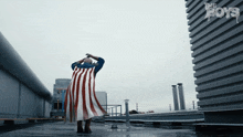 a man in an american flag cape stands on a rooftop in front of a sign that says " the boys "