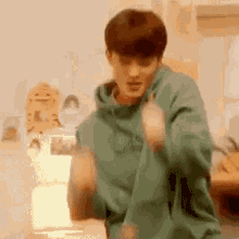 a man in a green hoodie is dancing in a room with a laptop .