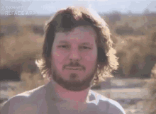 a man with long hair and a beard is standing in a field looking at the camera .