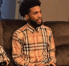 a man with a beard is sitting on a couch wearing a plaid shirt and holding a video game controller .