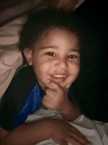 a little boy in a blue shirt is smiling and holding his hand to his chin