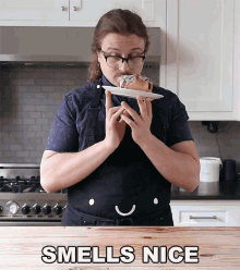 a woman in an apron is holding a plate with a donut on it and smelling it with the words smells nice below her