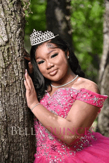 a woman in a pink dress and tiara leans against a tree trunk