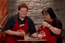 two women in red aprons are standing next to each other in front of a table with food on it .