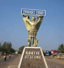 a statue of a man with wings holds a sign that says " kratie welcome "