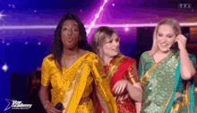 three women wearing saris are standing on a stage and one is holding a microphone .