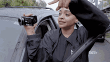 a woman in a black jacket is holding a camera in front of a car that says bet on the bottom