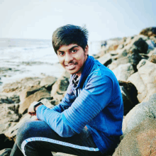 a young man in a blue jacket sits on a rocky shoreline