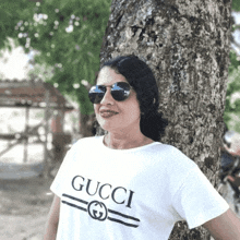 a woman wearing sunglasses and a white gucci t-shirt leans against a tree