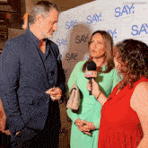 a man in a suit and a woman in a green dress are being interviewed by two women in front of a wall that says say