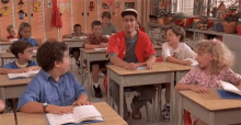 a group of children are sitting at their desks in a classroom with a man in a red shirt standing in the middle .