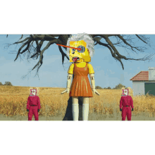 a yellow doll with a robot head stands in a field with two people in red suits