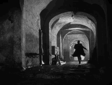 a black and white photo of a man in a trench coat walking through a dark tunnel