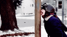 a young girl wearing a helmet and scarf is sticking her tongue out of a wooden post in the snow .