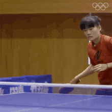 a girl in a red shirt is playing ping pong on a table with the olympic rings in the background