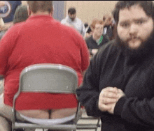 a man with a beard is standing next to a man in a red shirt who is sitting in a chair .