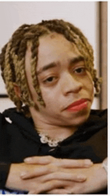 a young man with dreadlocks and red lipstick is making a funny face with his hands folded .