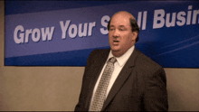 a bald man in a suit and tie stands in front of a sign that says grow your small business