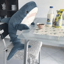 a stuffed shark is sitting on a chair next to a table with a bowl of cereal .