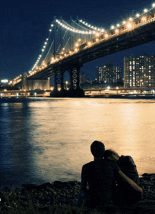 a couple sits on the shore of a river looking at a bridge at night