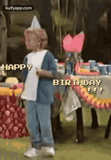 a young boy wearing a party hat is standing in front of a table with a birthday cake on it .