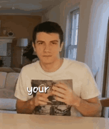 a young man in a white t-shirt is making a heart shape with his hands while sitting at a table .